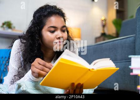 Nahaufnahme eines Mädchens, das vor dem Sofa Buch liest, indem es zu Hause auf dem Boden liegt - Konzept von Hobbys, Weisheit und Selbstentwicklung Stockfoto