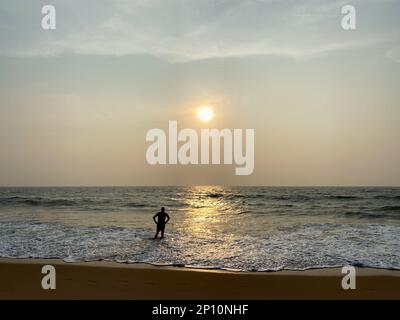 Candolim, Goa, Indien - Januar 2023: Die Silhouette eines Mannes, der den Sonnenuntergang genießt, während er im Wasser des Meeres steht. Stockfoto