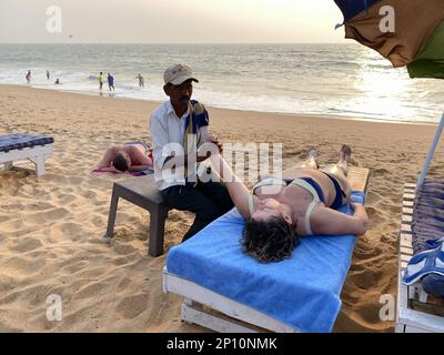 Candolim, Goa, Indien - Januar 2023: Ein indischer Mann, der einer Touristenfrau, die auf einem Strandbett am Strand von Goa liegt, eine Massage gibt. Stockfoto