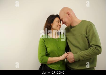 Ein Mann mittleren Alters, der erleichtert ist, die Hand seiner Frau zu halten, erzählt freudig die Geschichte im Ohr. Fesselnde Liebe, die auf weißem Hintergrund zieht, in identischen grünen Pullover, Mann und Frau flirten, während sie sich gegenseitig festhalten Stockfoto