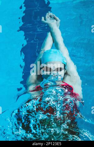 Draufsicht einer jungen Schwimmerin, die im Pool trainiert und auf ihrem Rücken unter Wasser schwimmt. Schwimmt unter Wasser und bläst Luftringe. Stockfoto