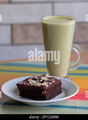 Tallglas mit veganem Milchmatcha-Tee auf Pflanzenbasis und veganem Schokoladenbrownie im Vordergrund. Serviert auf einer farbenfrohen Glasplatte. Stockfoto