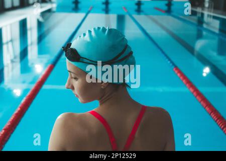Glückliche, muskuläre Schwimmerin mit Schutzbrille und Kappe im Pool und repräsentiert das Konzept von Gesundheit und guter Form. Schönes weibliches Model im Wasser Stockfoto