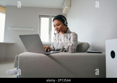 Eine Frau mit Headset auf dem Kopf sitzt auf dem Sofa mit einem Laptop Stockfoto