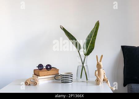 Stillleben mit einem Holzhasen auf Scharnieren im Innenraum. Eine Vase mit zwei grünen Palmenfronten, einem weiß-schwarz gestreiften Becher, einem Stapel Bücher und einem Woo Stockfoto