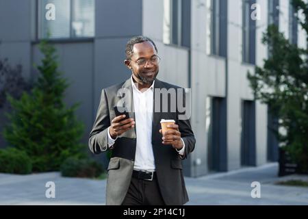 Kaffeepause. Ein erfolgreicher afroamerikanischer Geschäftsmann in einem Business-Anzug steht auf der Straße neben einem Wolkenkratzer in Anzug und Gläsern, hält eine Tasse Kaffee zum Mitnehmen und benutzt ein Handy. Stockfoto