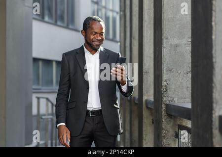 Ein erfolgreicher afroamerikanischer Geschäftsmann in einem Business-Anzug steht auf der Straße neben einem Wolkenkratzer, trägt einen Anzug und benutzt ein Handy. Stockfoto