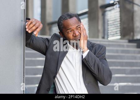Zahnschmerzen auf der Straße. Ein Afroamerikaner im Anzug steht vor einem Büro und hält seine Wange, leidet an Zahnschmerzen. Stockfoto