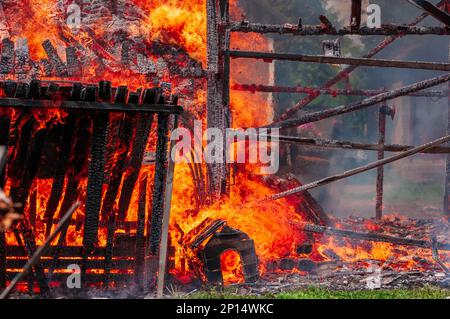 Die Holzscheune mit der hundehütte davor, die vollständig vom Feuer verschlungen wurde, wird bis auf den Boden abgebrannt. Nahaufnahme eines Feuerausbruchs aus einer brennenden Scheune. Stockfoto