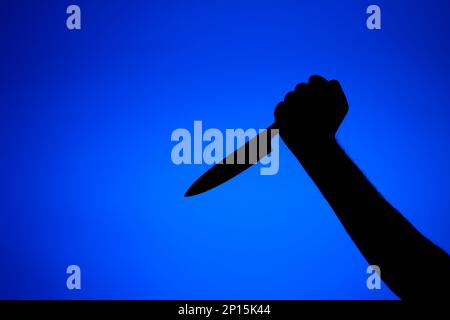 Silhouette auf einem männlichen Arm hält ein langes Küchenmesser bedrohlich. Nahaufnahme des Studios, isoliert auf blauem Hintergrund. Stockfoto