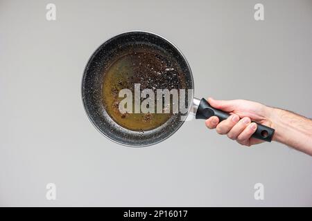 Weiße männliche Hand, die eine alte, mit braunem verbranntem Öl und auf grau isoliertem Fett befleckte Bratpfanne hält. Stockfoto