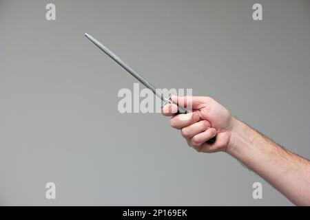 Kaukasische männliche Hand mit einem Schärfstab aus Stahlmesser isoliert auf grauem Hintergrund. Stockfoto