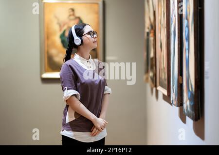 Seitenansicht einer jungen, hübschen weißen Frau, die Kopfhörer trägt und antike Artefakte betrachtet. Student besucht Kunstausstellung. Kulturbegriff Stockfoto