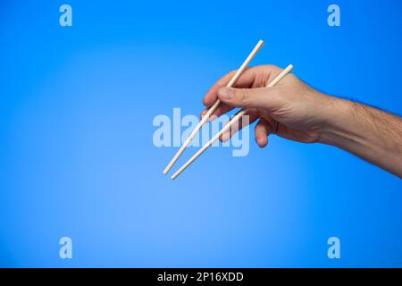 Asiatische Holzstäbchen in der Hand gehalten vom männlichen, kaukasischen Handstudio isoliert auf blauem Hintergrund. Stockfoto