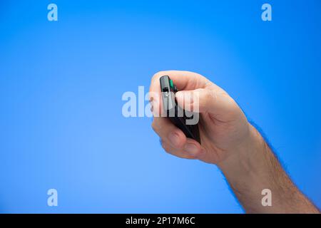 Weißer männlicher Handgriff, der Tasten auf einer digitalen Fernbedienung hält und drückt, die zur Kamera abgewinkelt ist, isoliert auf blau. Stockfoto