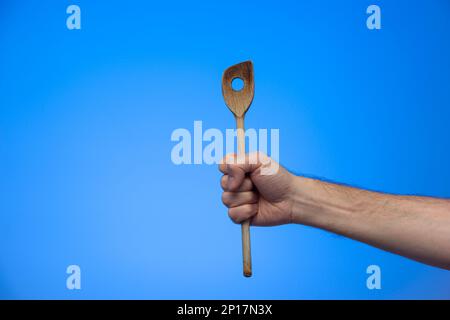 Weiße männliche Hand mit einem Holzlöffel Nahaufnahme isoliert auf blauem Hintergrund. Stockfoto