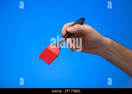 Kaukasischer männlicher Handgriff mit Silikon-roter und schwarzer Küchenbürste isoliert auf blauem Hintergrund. Stockfoto