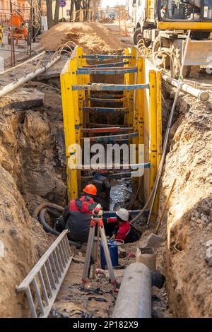 Hydraulikbagger füllt Tiefenaushub, unterstützt durch Grabenkasten mit Rohreinstreu Erbsenkies während der Installation des Entwässerungsrohrs. Rekonstruktion der Technik Stockfoto