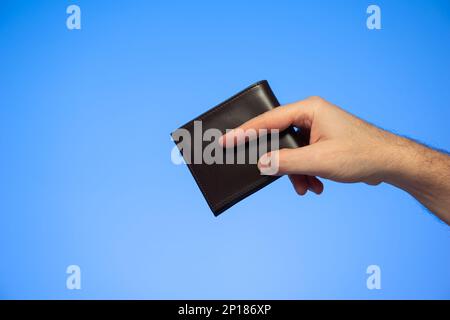 Weiße männliche Hand mit schwarzer Lederbörse, die ein Studio mit Gesten auf Blau isoliert macht. Stockfoto
