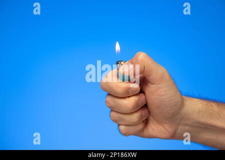Weiße männliche Hand mit einem beleuchteten manuellen Feuerzeug-Studio, isoliert auf blau. Stockfoto