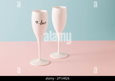 Zwei Champagnergläser auf einer pinkfarbenen und blauen Oberfläche mit der Aufschrift „Love“ auf einer der Gläser Stockfoto