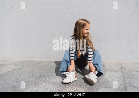 Eine lange, lächelnde Frau in Jeansweste und blaue Jeans, die auf dem Basketball neben der Wand sitzt, Stockbild Stockfoto