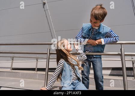 Fröhliche und gut gekleidete Kinder in legerer Jeanskleidung, die sich neben dem Gebäude mit Metallgeländern posieren, Stockbild Stockfoto