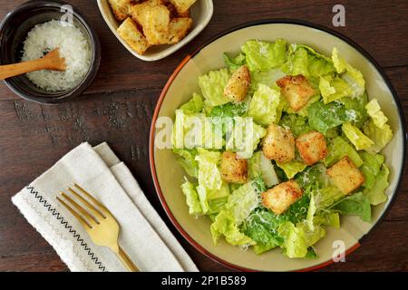 Frischer und gesunder caesar-Salat mit Römersalat, geriebenem Parmesankäse und hausgemachten Croutons mit Goldgabel auf dunklem Holzhintergrund von oben. Stockfoto
