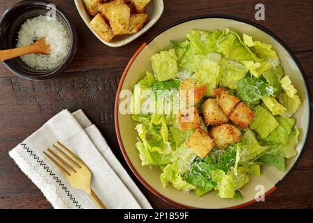 Frischer und gesunder caesar-Salat mit Römersalat, geriebenem Parmesankäse und hausgemachten Croutons mit Goldgabel auf dunklem Holzhintergrund von oben. Stockfoto