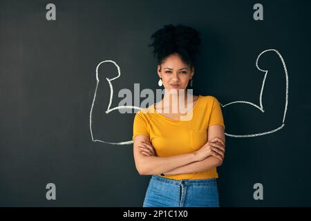 Ich bin eine starke Frau. Aufnahme einer Frau, die mit einer Kreidedarstellung von sich biegenden Muskeln vor dunklem Hintergrund posiert. Stockfoto