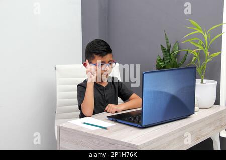 Ein 7-jähriger Latino-Junge mit Brille, der zu Hause zu Hause an Online-Kursen teilnimmt, ist überrascht, dass er mit einem Laptop und dem Studium an einem Tisch sitzt Stockfoto