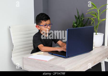 Ein 7-jähriger Latino-Junge mit Brille, der zu Hause zu Hause an Online-Kursen teilnimmt, ist überrascht, dass er mit einem Laptop und dem Studium an einem Tisch sitzt Stockfoto