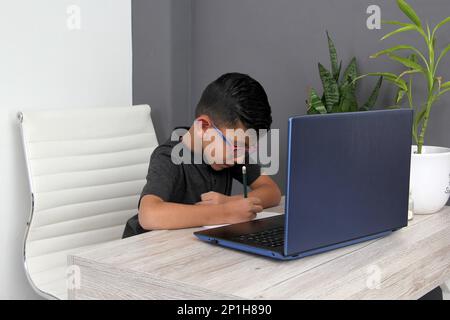 Ein 7-jähriger Latino-Junge mit Brille, der zu Hause zu Hause an Online-Kursen teilnimmt, ist überrascht, dass er mit einem Laptop und dem Studium an einem Tisch sitzt Stockfoto
