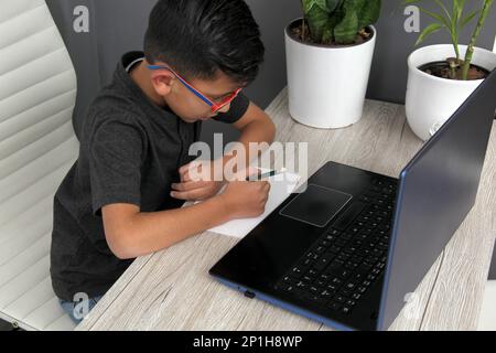 Ein 7-jähriger Latino-Junge mit Brille, der zu Hause zu Hause an Online-Kursen teilnimmt, ist überrascht, dass er mit einem Laptop und dem Studium an einem Tisch sitzt Stockfoto