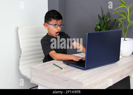Ein 7-jähriger Latino-Junge mit Brille, der zu Hause zu Hause an Online-Kursen teilnimmt, ist überrascht, dass er mit einem Laptop und dem Studium an einem Tisch sitzt Stockfoto