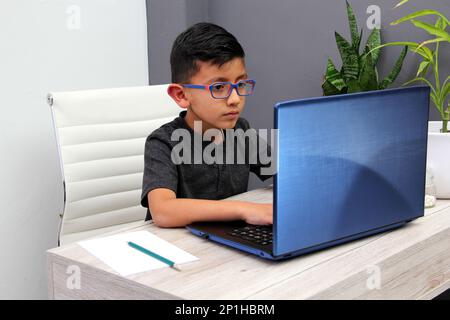 Ein 7-jähriger Latino-Junge mit Brille, der zu Hause zu Hause an Online-Kursen teilnimmt, ist überrascht, dass er mit einem Laptop und dem Studium an einem Tisch sitzt Stockfoto