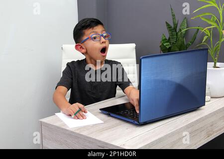 Ein 7-jähriger Latino-Junge mit Brille, der zu Hause zu Hause an Online-Kursen teilnimmt, ist überrascht, dass er mit einem Laptop und dem Studium an einem Tisch sitzt Stockfoto