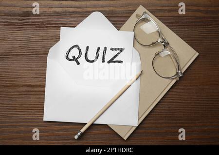 Papier mit Wörtern QUIZ in Umschlag, Bleistift, Notizbuch und Brille auf Holztisch, flach liegend Stockfoto