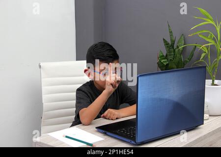 Ein 7-jähriger Latino-Junge mit Brille, der zu Hause zu Hause an Online-Kursen teilnimmt, ist überrascht, dass er mit einem Laptop und dem Studium an einem Tisch sitzt Stockfoto