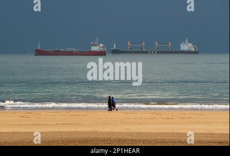 Menschen, die am Strand spazieren gehen, mit vor Anker liegenden Schiffen, die an einem Wintermorgen auf den Hafen von Santander Cantabria Spanien warten Stockfoto