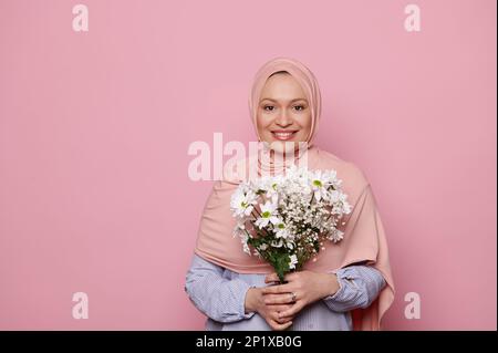 Wunderschöne muslimische Frau in pinkem Hijab, hält einen Strauß Kamillenblüten, lächelt in die Kamera, isolierter rosa Hintergrund Stockfoto