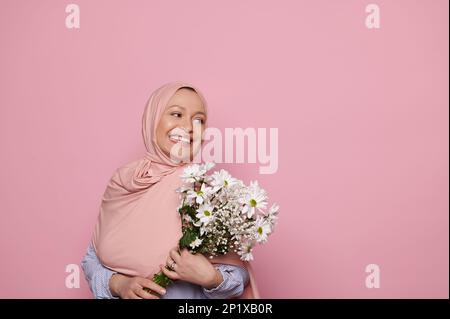 Wunderschöne muslimische Frau in pinkfarbenem Hijab, mit einem Haufen weißer Wildblumen, lächelnd in die Werbefläche, isolierter Hintergrund Stockfoto