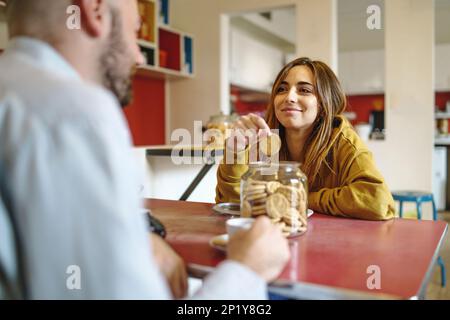 Ein Paar, das zusammen an einem Tisch frühstückt, die Frau nimmt einen Keks aus einem Glas, während sie ihren Partner anschaut, der Kaffee trinkt - Menschen leben Stockfoto