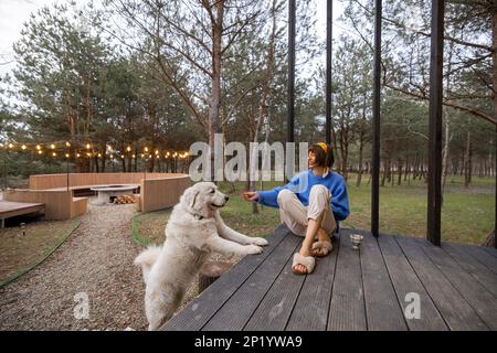 Eine Frau mit Hund ruht in der Nähe einer hölzernen Hütte in der Natur Stockfoto