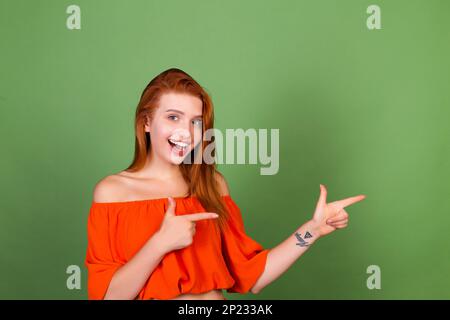 Junge Frau mit rotem Haar in lässiger orangefarbener Bluse auf grünem Hintergrund lächelt und lacht rechts Stockfoto