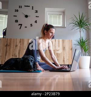 Junge Frau, die auf dem Boden eines Wohnzimmers sitzt und einen Laptop benutzt oder daran arbeitet. Ihr schwarzer labrador Retriever Hund liegt neben ihr auf einer Matte. Stockfoto
