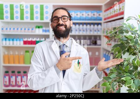 Hispanic Mann mit Bart arbeitet in der Apotheke Drogerie erstaunt und lächelt zur Kamera, während mit der Hand und zeigen mit dem Finger. Stockfoto