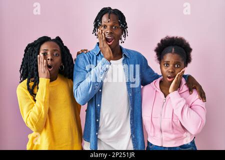 Eine Gruppe von drei jungen Schwarzen, die zusammen über einem pinkfarbenen Hintergrund stehen, ängstlich und schockiert sind, überrascht sind und einen überraschenden Gesichtsausdruck mit Händen im Gesicht haben Stockfoto
