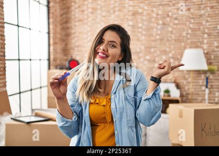 Junge hispanische Frau, die das Ergebnis des Schwangerschaftstests in der neuen Wohnung hält und glücklich lächelt, mit Hand und Finger zur Seite zeigt Stockfoto