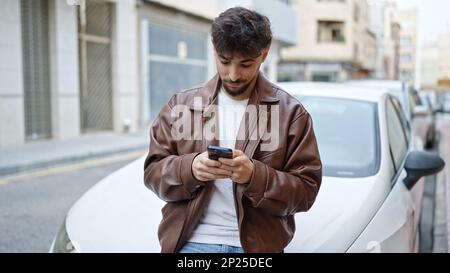 Ein junger araber, der sein Smartphone benutzt und sich auf der Straße an ein Auto lehnt Stockfoto